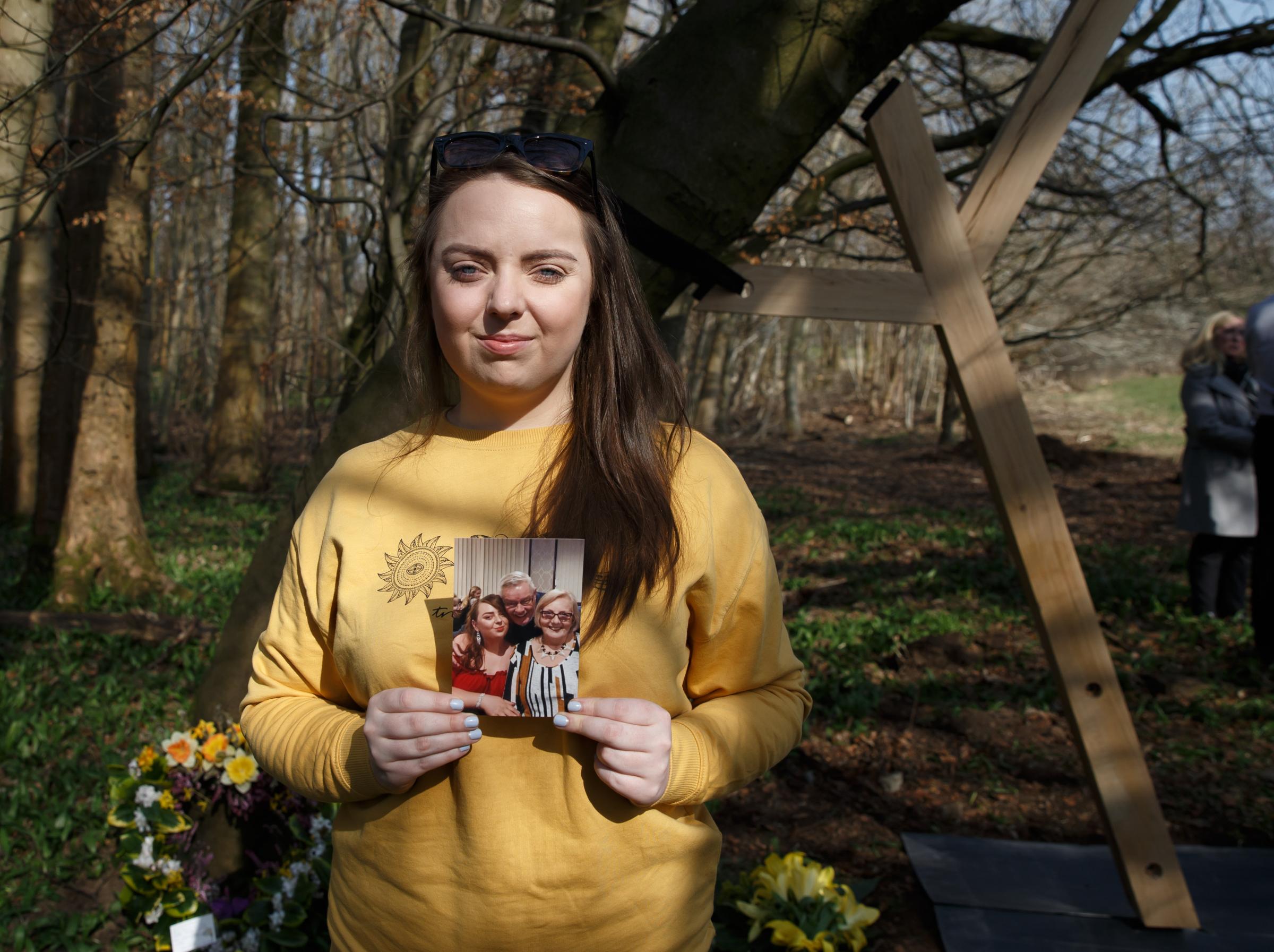 Keren Nairns pictured at Pollok Country Park. She lost her grandparents, Agnes Addison and David Wilson to Covid. Next to the floral tributes is the first of a series of wooden tree supports created by The Heralds covid memorial artist Alec Finlay.