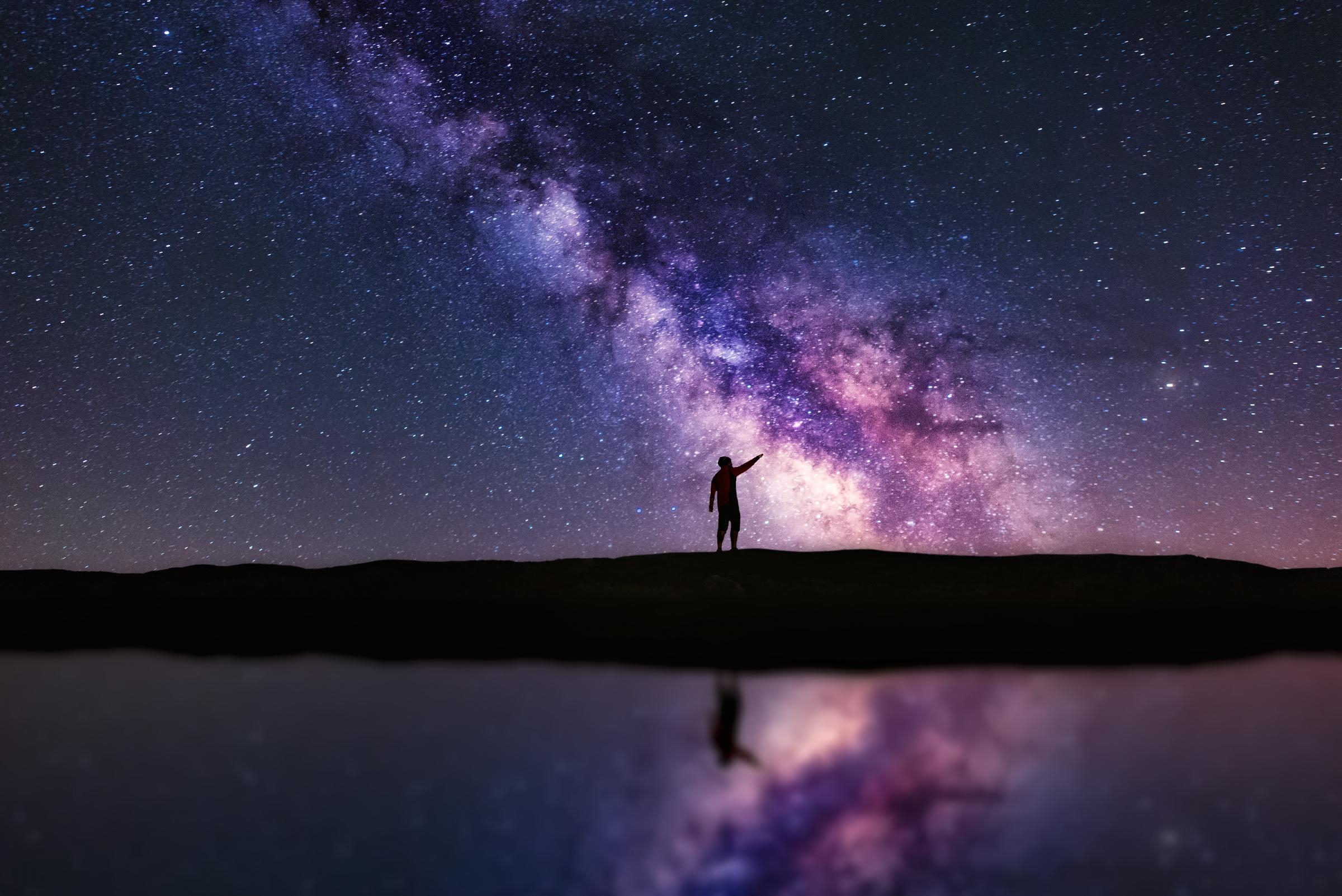 Person pointing up the Milky Way galaxy at night. The scene is reflected in water. A Veiga, Ourense, Spain.